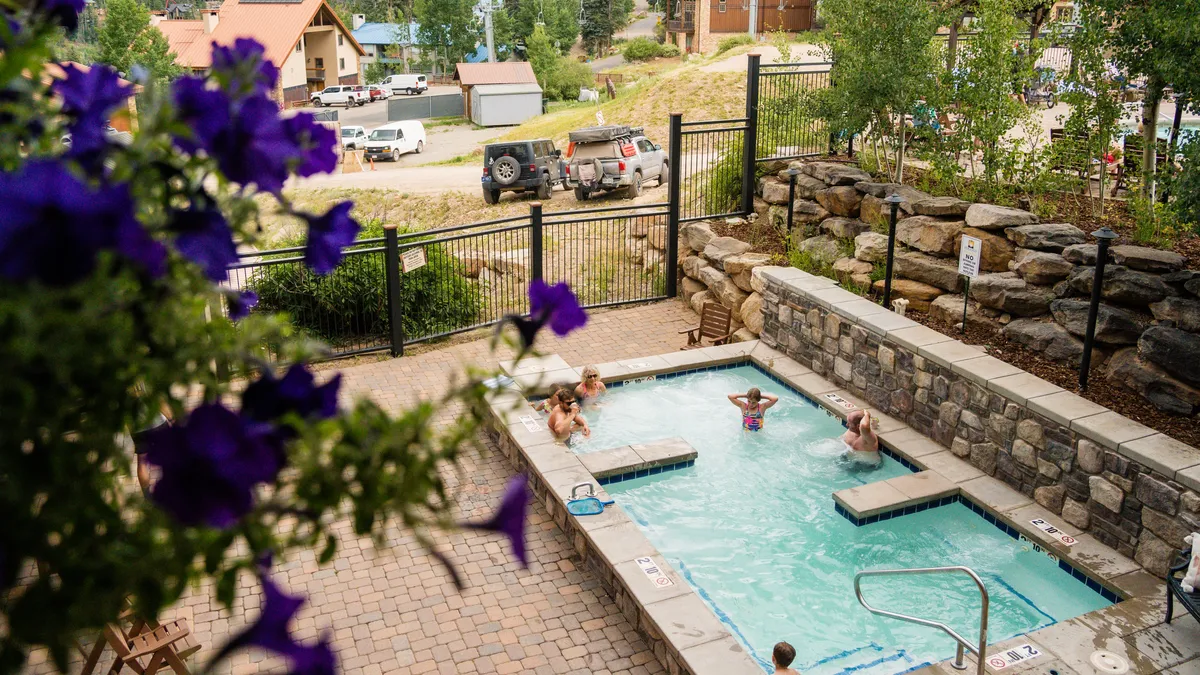 A cheerful exterior shot of a swimming pool surrounded by purple flowers and a stone wall. The scene is vibrant, with children and families likely gathered for swimming, sunbathing, and socializing, evoking a sense of joy and leisure.