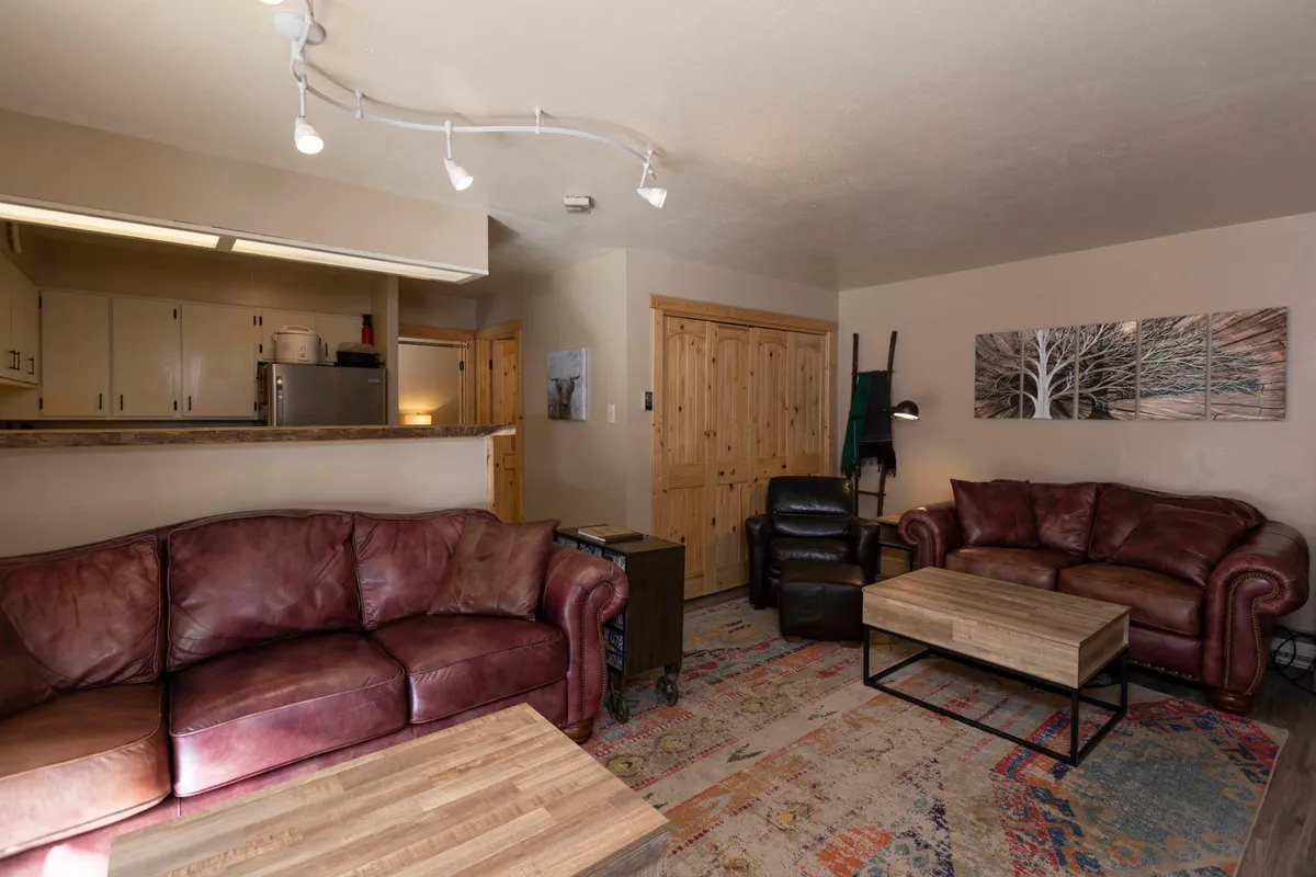 A cozy and inviting living area furnished with plush leather sofas, a wooden coffee table, and tasteful wall art, seamlessly connected to a kitchen bar. The space is designed for relaxation and socializing, providing a comfortable atmosphere ideal for families or friends after a day on the slopes.