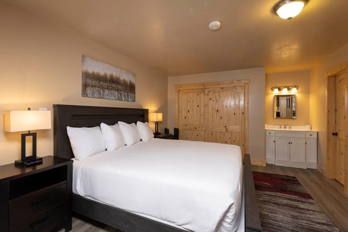 The image showcases a cozy bedroom with a plush king bed, inviting nightstands, soft lighting, and tasteful artwork. There is a glimpse of the nearby bathroom area, suggesting convenience and comfort. The overall atmosphere is warm and relaxing, perfect for unwinding after a day on the slopes.
