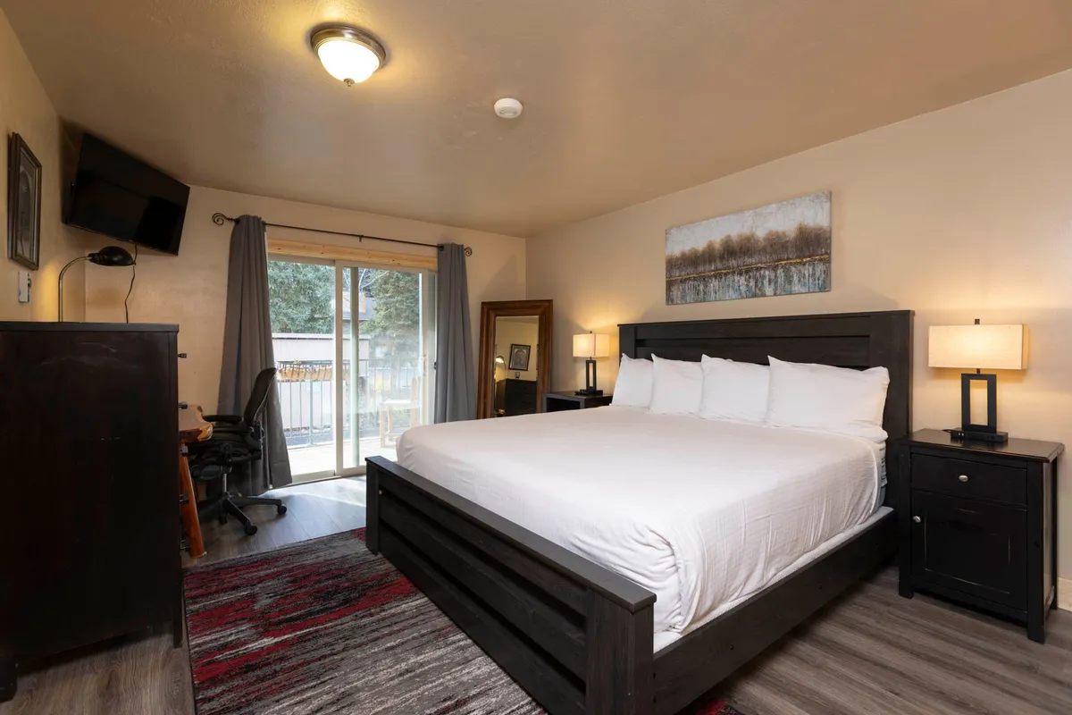 This image showcases a serene bedroom with a king-size bed centrally positioned, flanked by nightstands with lamps. A large window dressed with curtains allows for an abundance of natural light, and tasteful artwork adorns the wall. The space is thoughtfully arranged to maximize comfort and relaxation, with a small desk area visible for quiet tasks or reading.