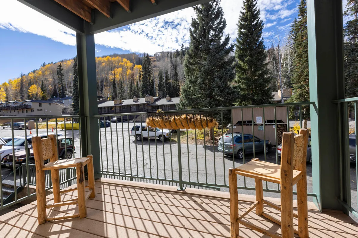 An inviting scene featuring two wooden chairs on a patio or balcony, overlooking a lush, forested hillside beneath a clear blue sky. The setting encourages relaxation and togetherness while enjoying the mountain air and natural surroundings.