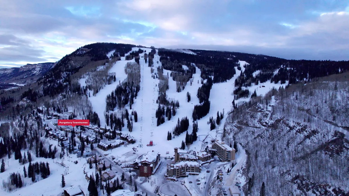 A sweeping winter view captures the Purgatory Townhomes nestled at the base of snow-blanketed ski slopes. The scene includes the townhomes in the foreground with the surrounding forest and mountains creating a dramatic, scenic backdrop. Tracks in the snow lead directly from the accommodations to the ski runs, illustrating immediate access to the slopes.