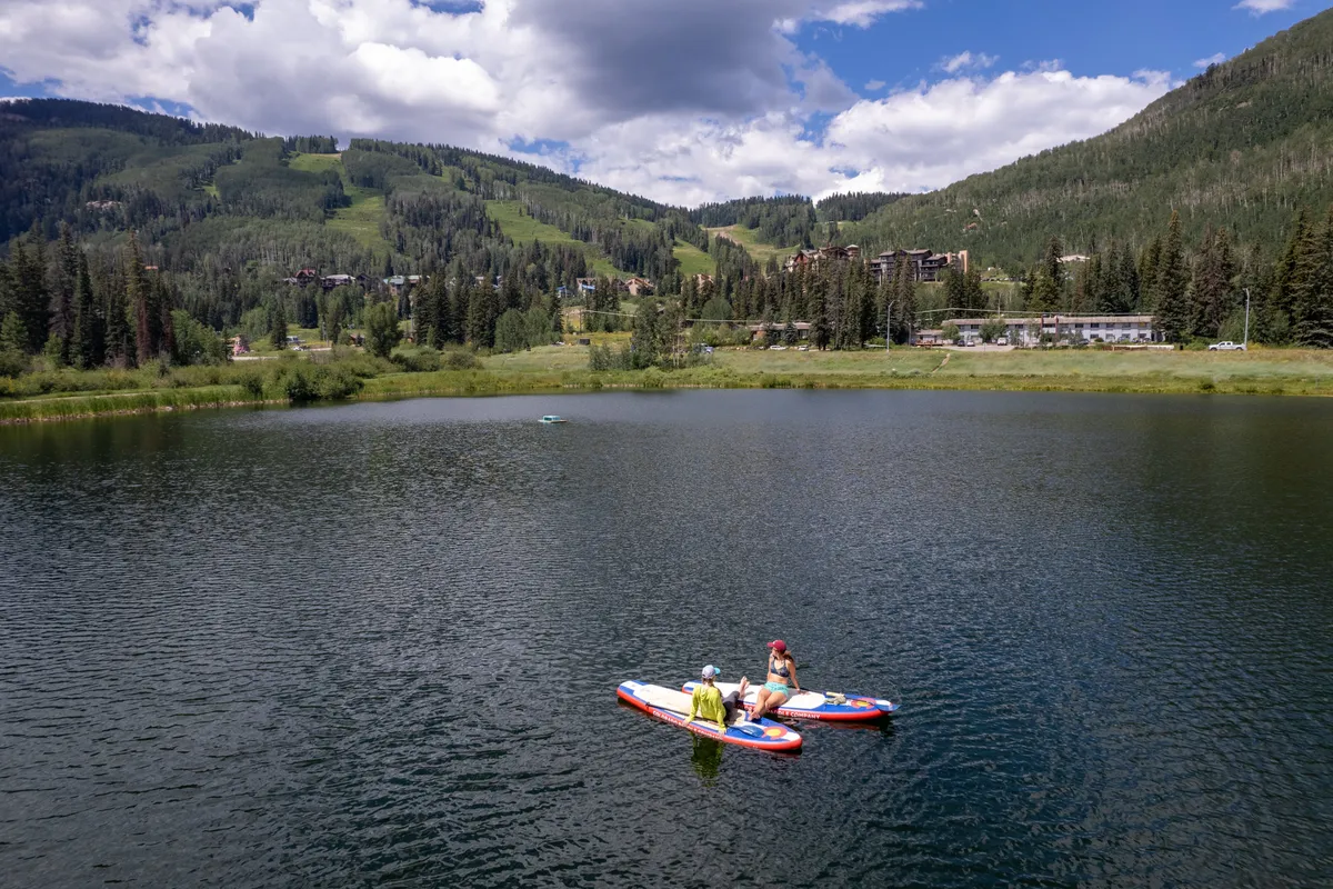 This image captures two people paddleboarding on a calm mountain lake at sunset, surrounded by a dramatic mountain backdrop. The tranquil water and expansive natural scenery evoke a sense of adventure and romantic escape.