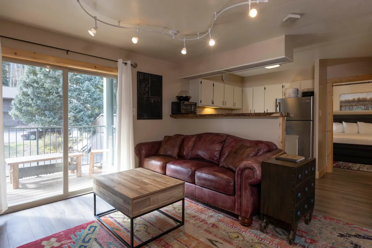 A warm, inviting living room with a brown leather sofa, a central coffee table, and a sliding glass door that lets in abundant natural light. Sunshine streams in, brightening the plush seating area and offering a glimpse of the scenery outside. The space feels cozy and perfect for relaxing or entertaining after a day on the slopes.