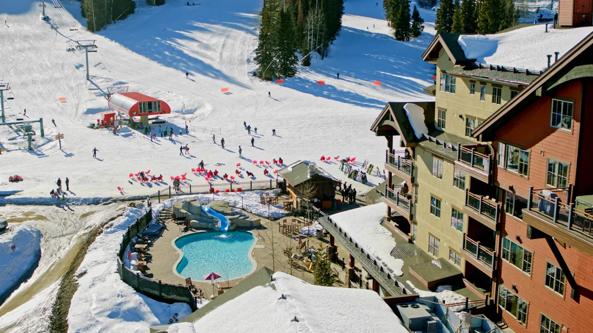 A lively exterior shot showing skiers in action on the slopes, the resort’s pool area, skiers walking nearby, and restaurants with the lodge facade in the background. The snowy scene is energetic, illustrating both active skiing and the relaxing après-ski options available just steps from the action.