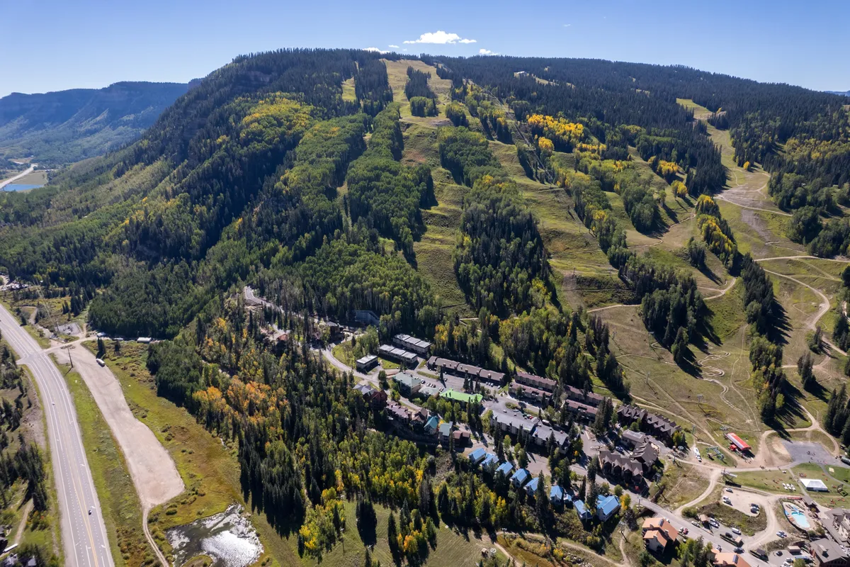 This exterior image captures sweeping views of mountain slopes bordered by a vibrant, colorful tree line. Residential buildings are nestled in the landscape, and the scene includes a roadway leading into the community. The overall atmosphere is lively but peaceful, suggesting opportunities for hiking, skiing, mountain biking, and sightseeing throughout the changing seasons.