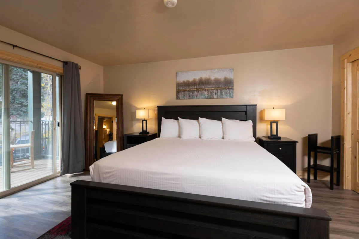 The photo captures a spacious bedroom with a large bed dressed in white linens, flanked by two bedside lamps. There's a wooden mirror and tasteful wall art, and a sliding glass door opens to a balcony, suggesting a peaceful transition between indoor rest and the outdoors. The overall mood is calm and cozy, ideal for reading or morning routines.