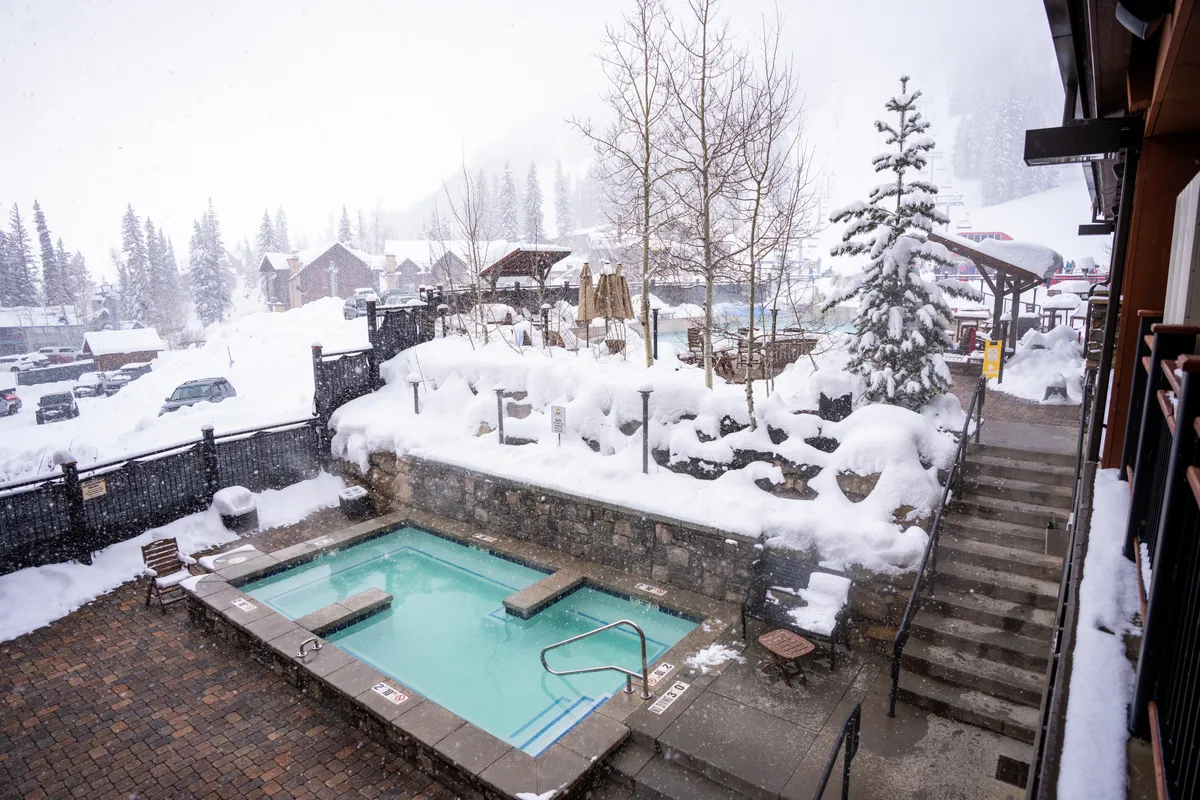 An inviting outdoor hot tub nestled among snow-covered trees and stone walls, with mountain vistas in the background. The setting exudes warmth and tranquility, perfect for relaxation after an adventurous day.