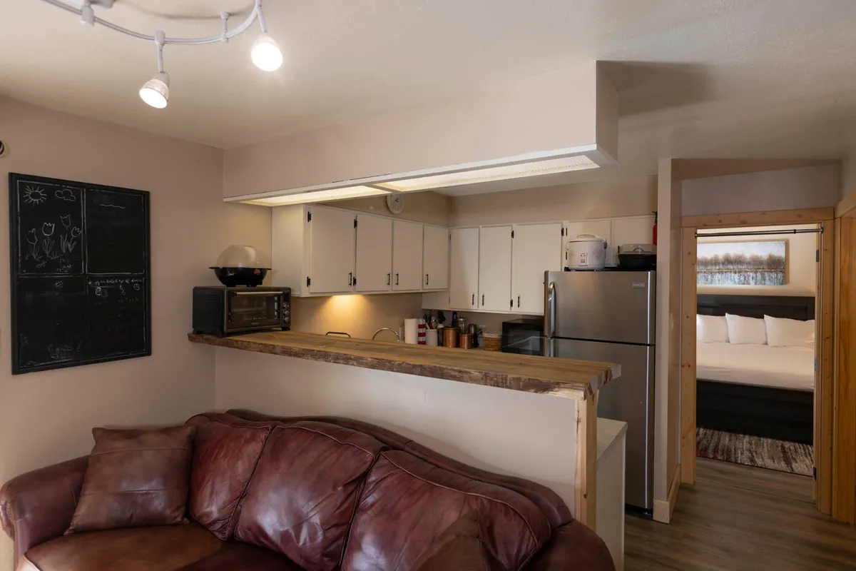 This interior scene features a leather couch, a kitchen counter, a blackboard, and a refrigerator. The space merges the kitchen with a casual seating area, highlighting a setting where cooking, relaxing, and socializing naturally blend together.